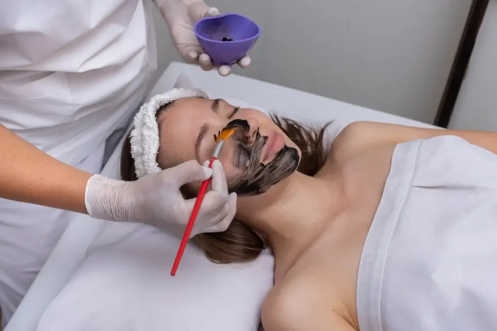 Practitioner applying a dark facial mask to a client’s face with a brush.