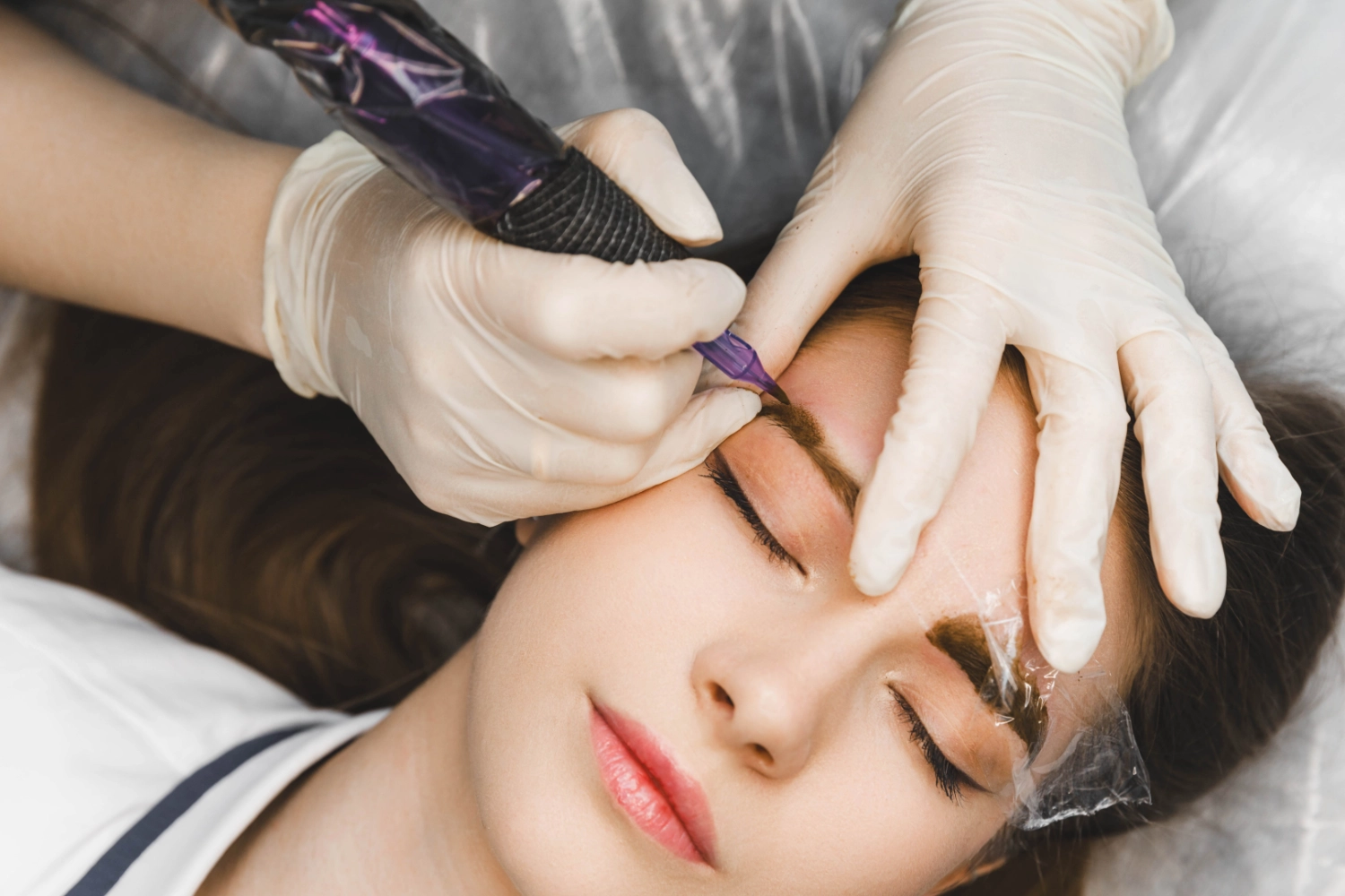 Practitioner using a handheld cosmetic tattoo device to enhance a client’s eyebrow.